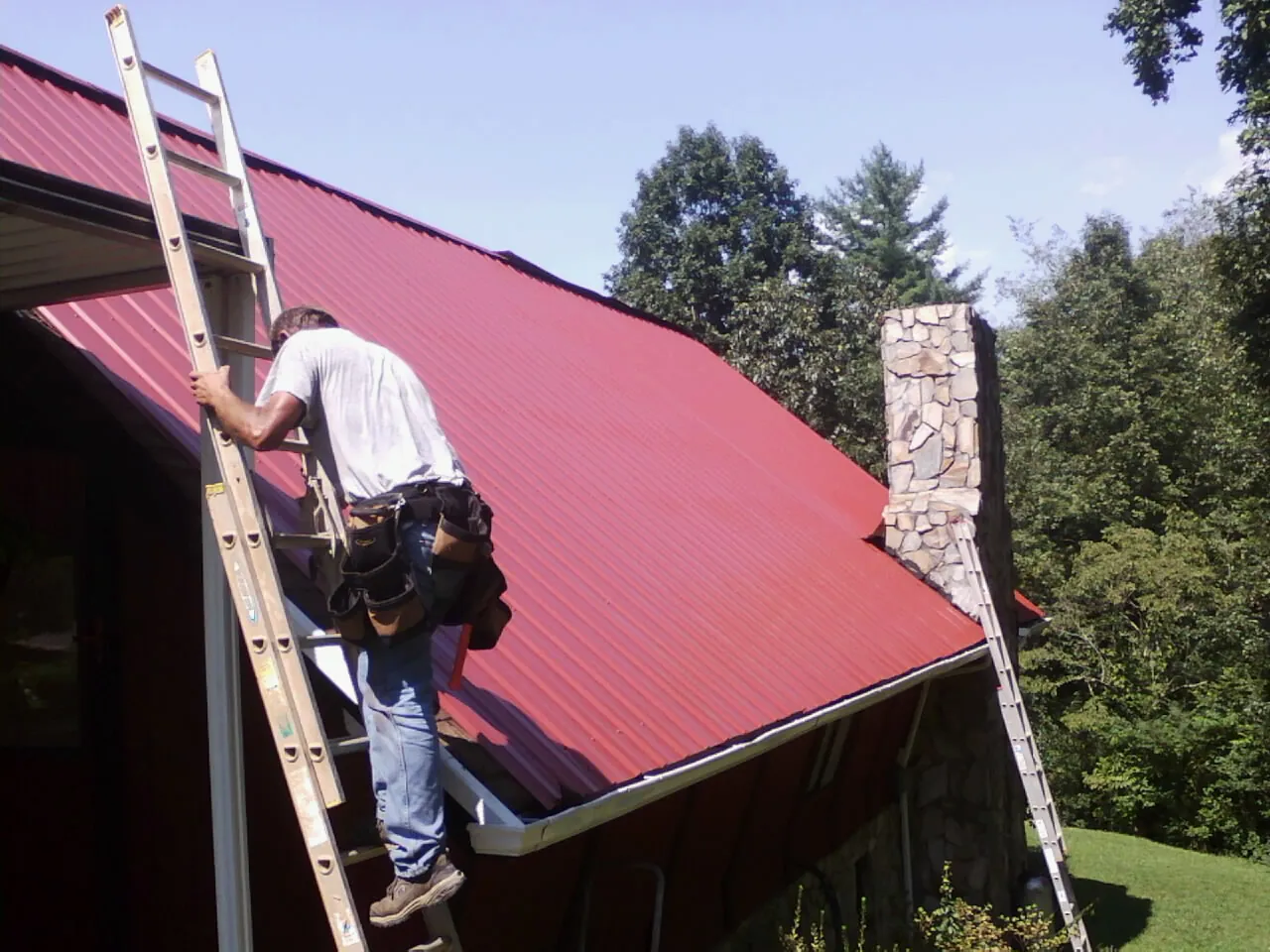 Roofer on a ladder beside a red metal roof and stone chimney — Soffit & Fascia Repair by Roofing Brothers in Ellicott City